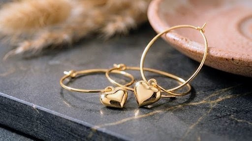 Deux créoles dorées à pendentif cœur posées sur une ardoise veinée d'or, près d'une coupelle en grès